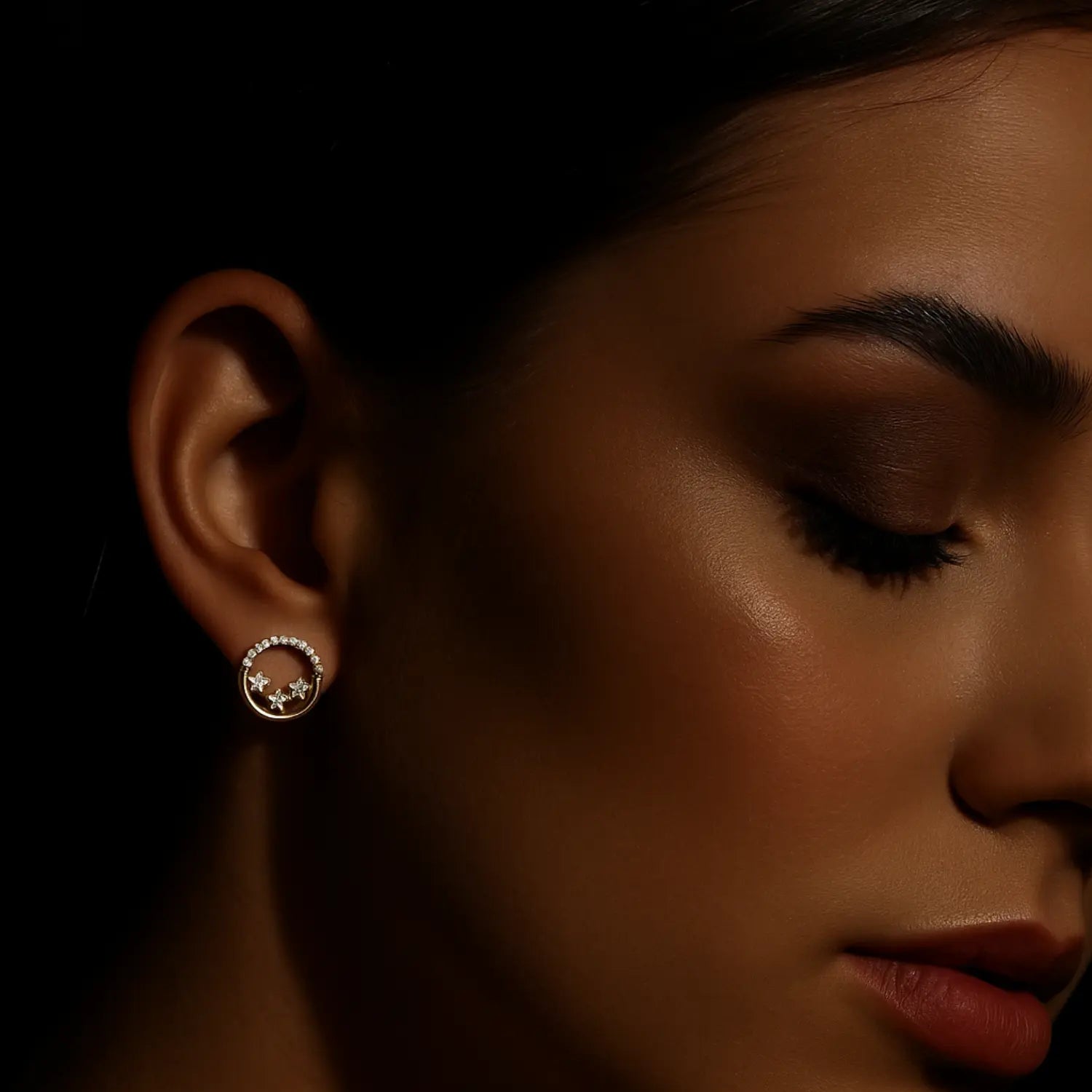 Zlaté kruhové náušnice s drobnými kamienkami na uchu ženy, tmavá produktová fotografia kolekcie